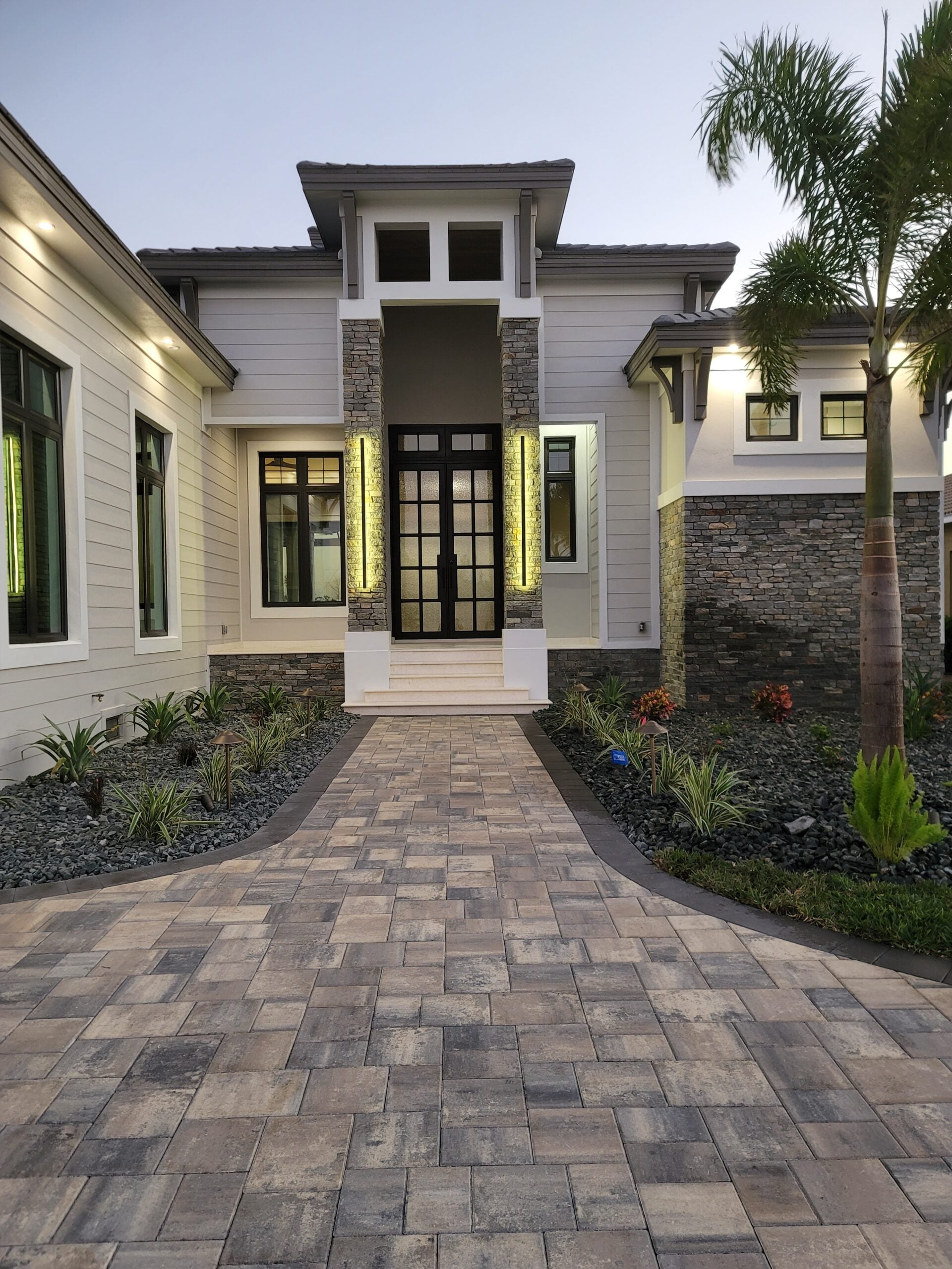 Modern home entry at dusk with illuminated stone columns and paver walkway